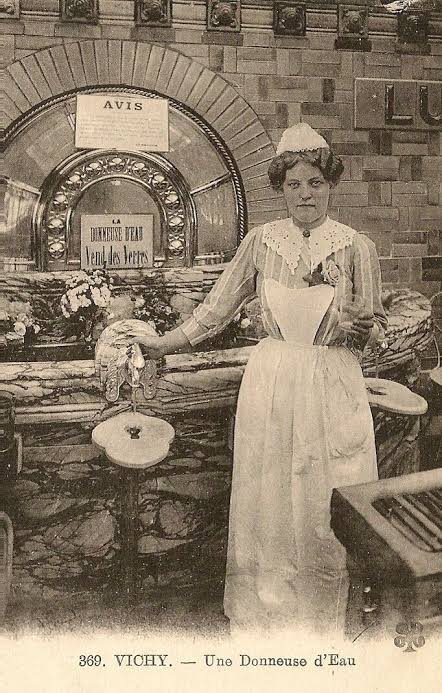 Carte postale ancienne légendée « Vichy — Une donneuse d'eau » : une femme en tablier blanc devant son comptoir de source