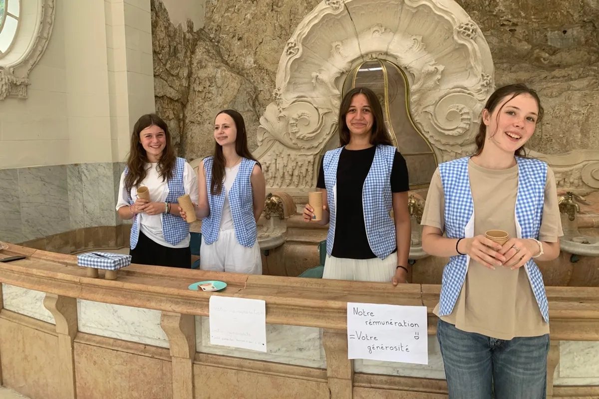 Quatre jeunes donneuses d'eaux en gilet vichy bleu et blanc, gobelets en main, derrière la balustrade de marbre du hall des sources