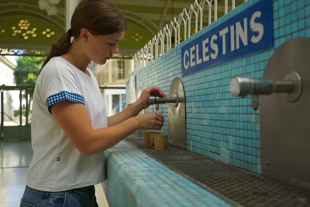 Au comptoir carrelé de bleu de la source des Célestins, une jeune femme remplit des gobelets au robinet
